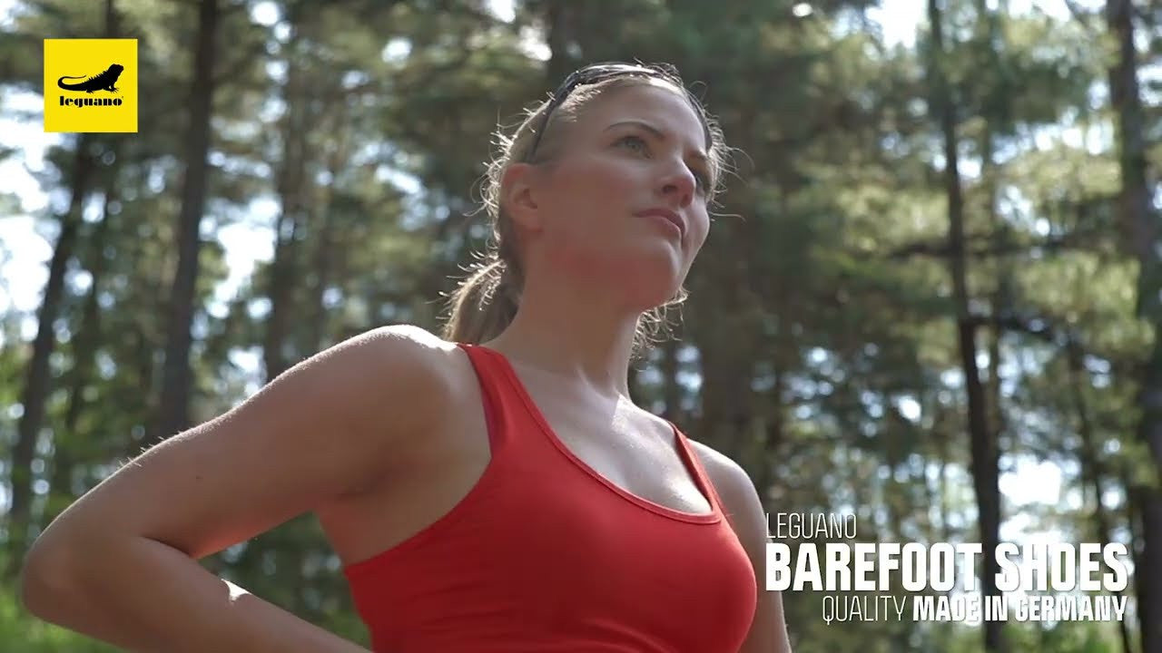 Thumbnail: Frau mit hochgeklappter Sonnenbrille in rotem Sporttop trägt Leguano Barfußschuhe im Wald. Logo schwarz-gelb oben links.