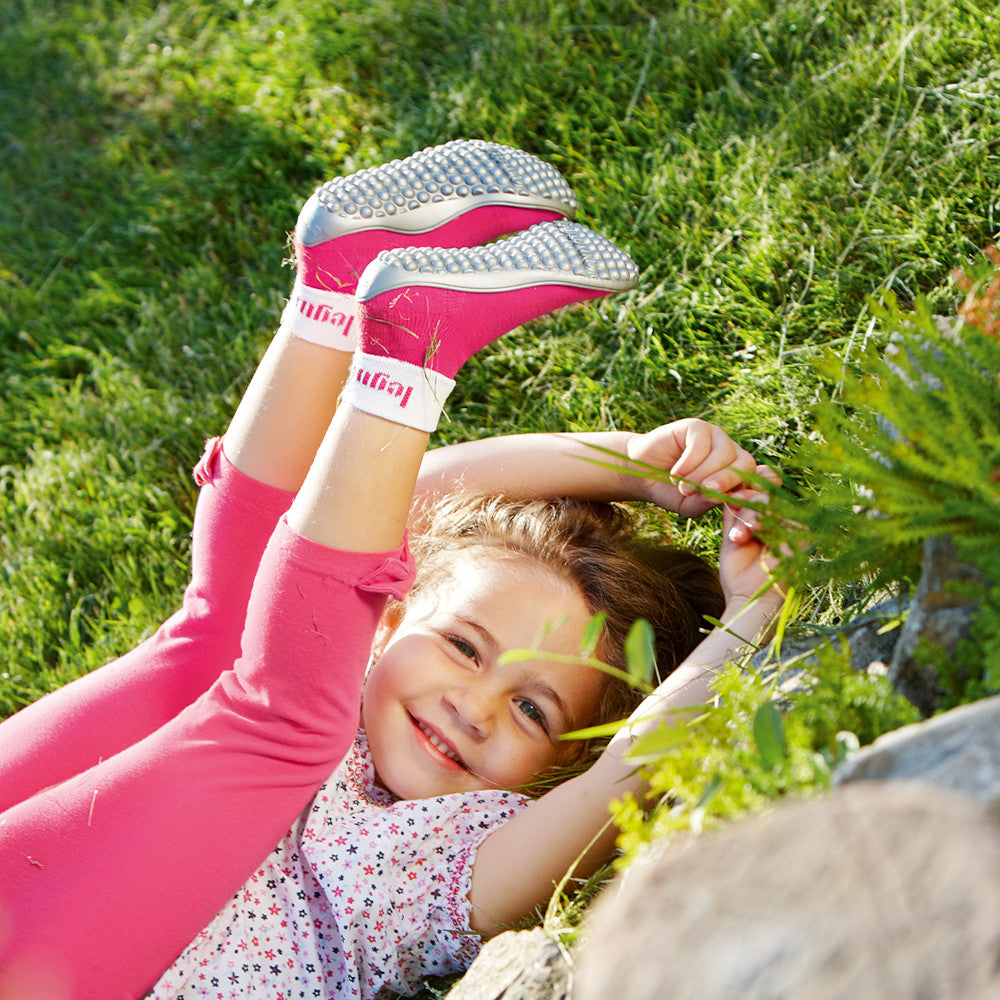 Leguano Barfußschuhe Kinder: Mädchen in pinken Barfußschuhen, lächelnd im Gras, Füße zum Himmel gestreckt